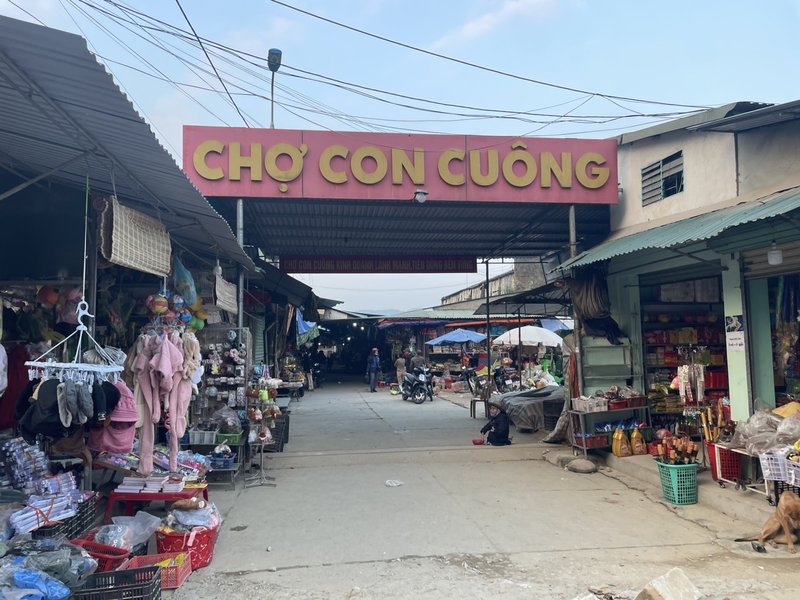 Bảng giá đất Nghệ An 2026 mới nhất Cổng chợ Con Cuông với biển chữ vàng nền đỏ, hai bên là các sạp hàng tạp hóa